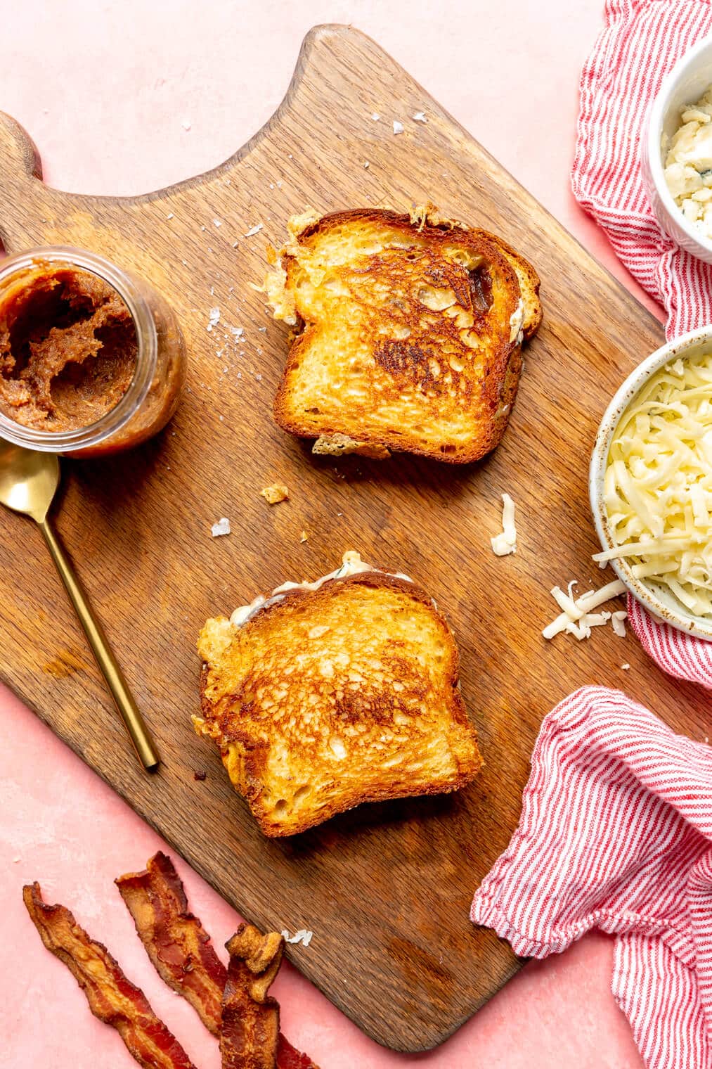 Two grilled cheese sandwiches on a wooden cutting board with a jar of bacon jam, bowl of sauerkraut, and slices of bacon.