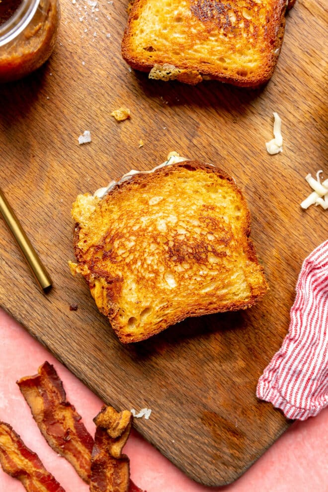 Grilled cheese sandwich on a wooden cutting board with a red and white striped linen and slices of bacon.
