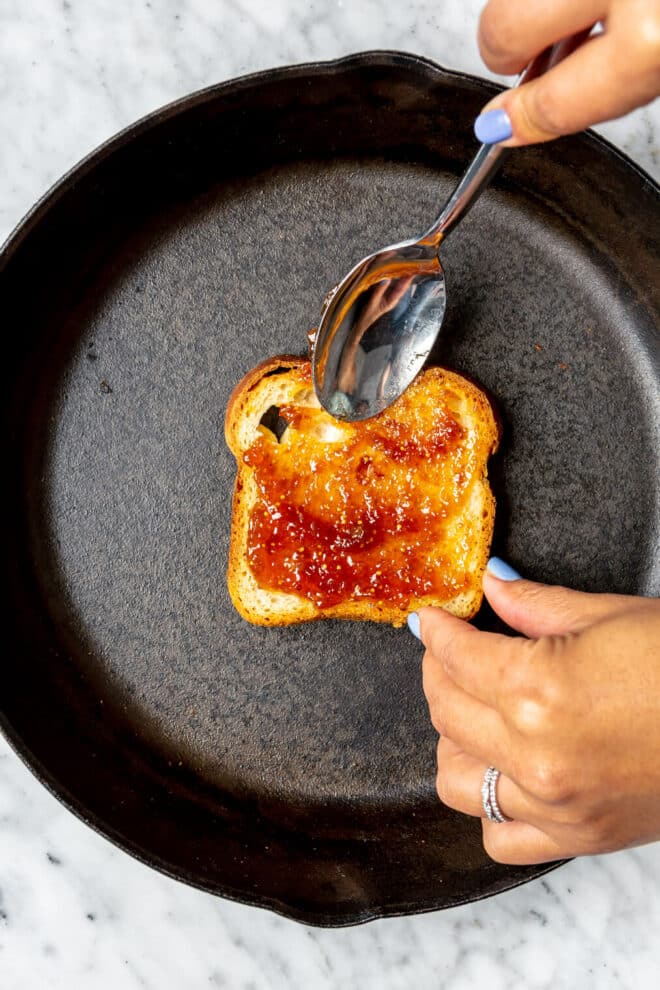 Hand holding spoon spreading fig jam on top of a slice of bread in a cast iron pan.
