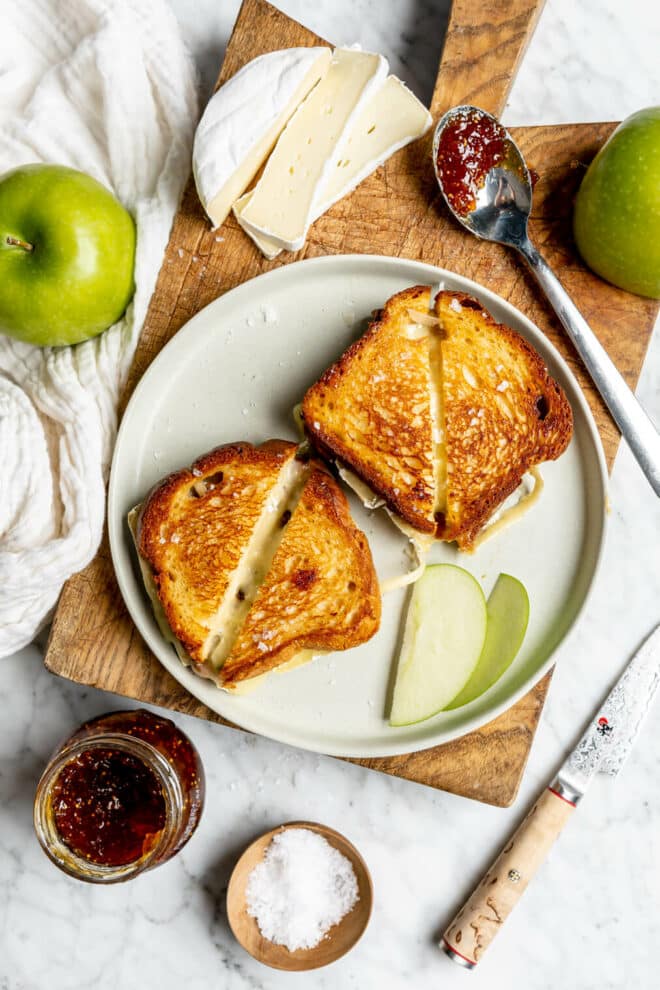 Two grilled cheese sandwiches on a round plate sitting on a wooden cutting board with sliced apples and a spoon with fig jam.