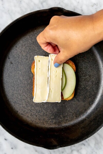 Hand layering sliced brie cheese on top of apples on a slice of bread in a cast iron pan.