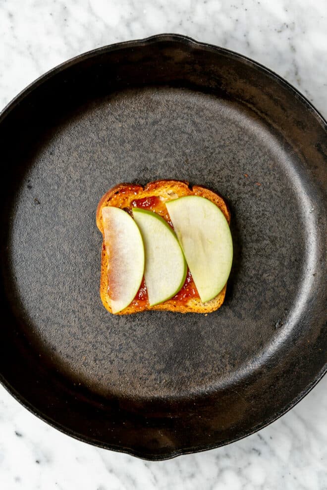 Three slices of apples on top of fig jam on a slice of bread in a cast iron pan.