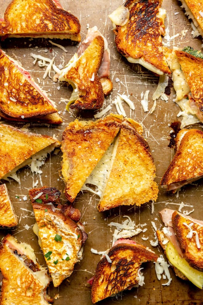 Grilled cheese sandwich halves and quarters on a brown baking sheet with cheese stretched and sprinkled around the sandwiches.