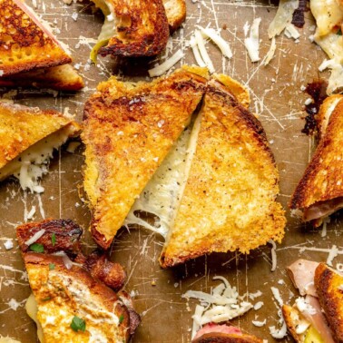 Grilled cheese sandwich halves and quarters on a brown baking sheet with cheese stretched and sprinkled around the sandwiches.
