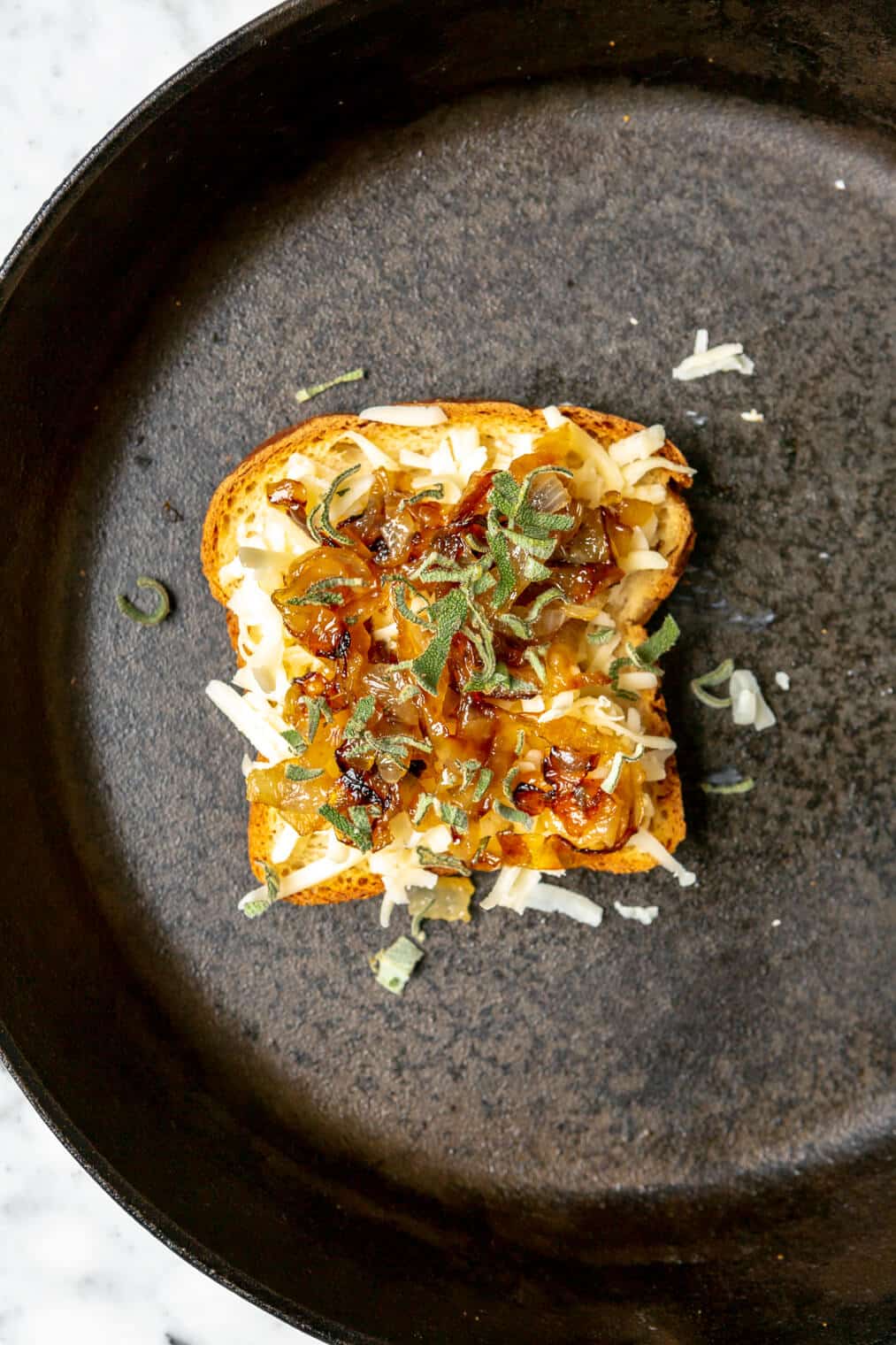 Slice of bread with shredded cheese, caramelized onions, and sage on top in a cast iron skillet.