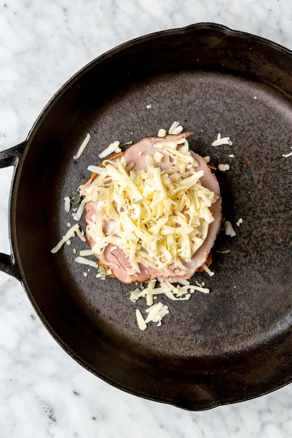 Cheese on top of ham on a slice of bread in a cast iron skillet.