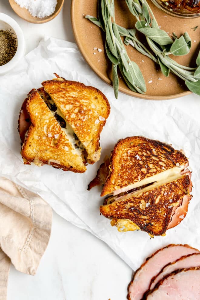 Two grilled cheese sandwiches on a white piece of parchment paper with ham, sage leaves, and cheese in the background.