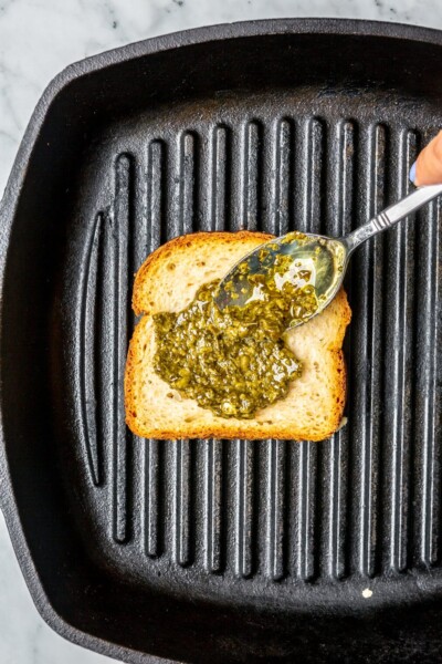 Spoon spreading layer of pesto sauce on top of a slice of bread in a cast iron grill pan.
