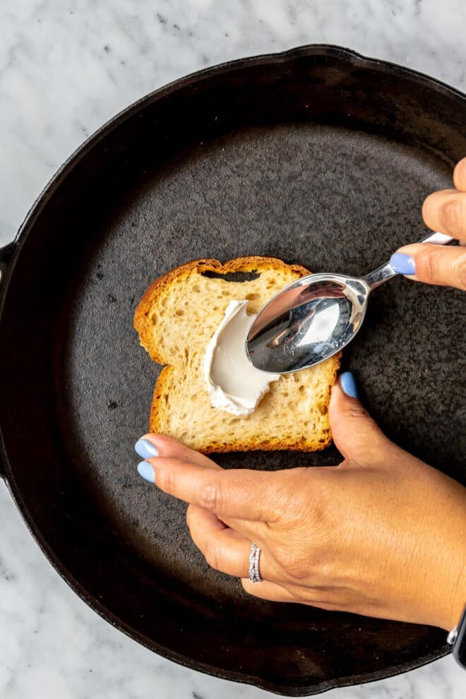 Hands holding a slice of bread and spoon spreading cream cheese on top of the bread in a cast iron skillet.