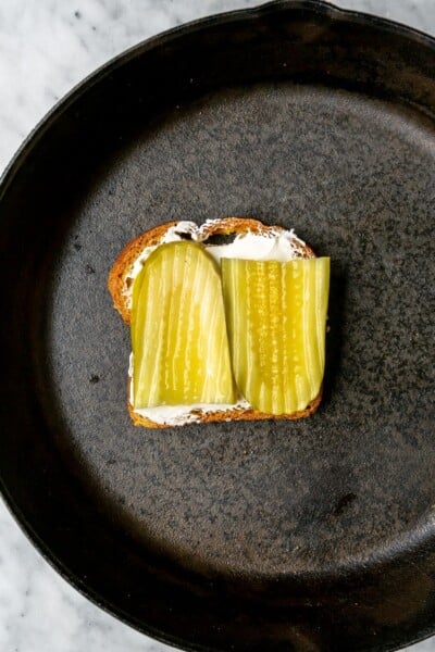 Two slices of pickle on top of cream cheese on a slice of bread in a cast iron skillet.