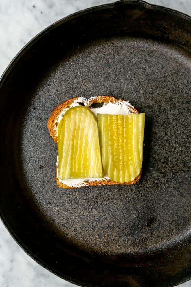 Two slices of pickle on top of cream cheese on a slice of bread in a cast iron skillet.