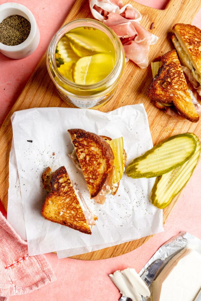 Sliced grilled cheese on a white piece of parchment paper with a jar of pickles and two slices of pickle next to it on a wooden cutting board.
