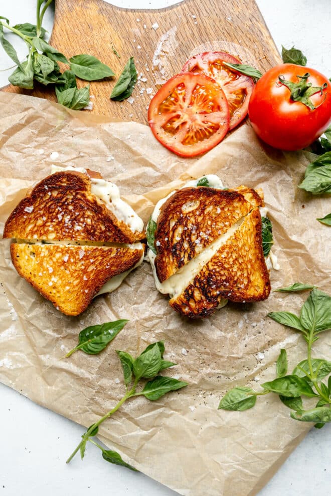 Two grilled cheese sandwiches on a brown piece of parchment paper with sliced tomato and basil leaves.