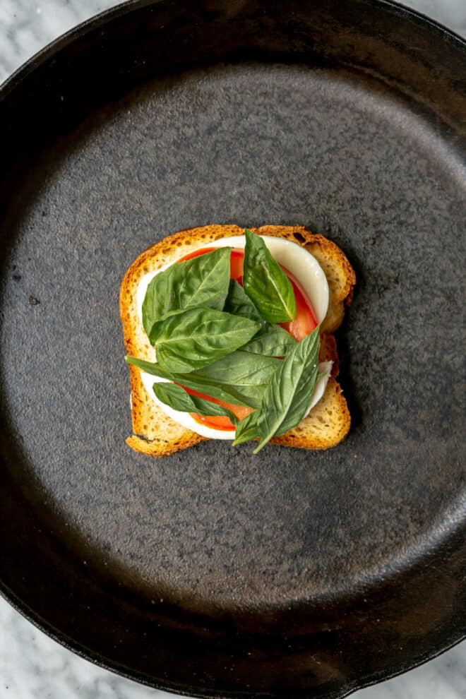 Basil leaves on top of a slice of tomato and mozzarella on a piece of bread in a cast iron skillet.