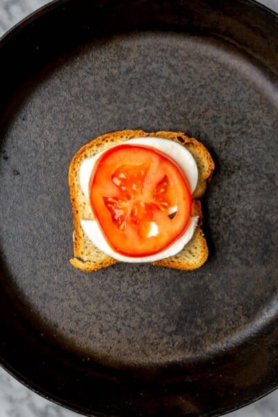 Slice of tomato layered on top of mozzarella on a piece of bread in a cast iron skillet.