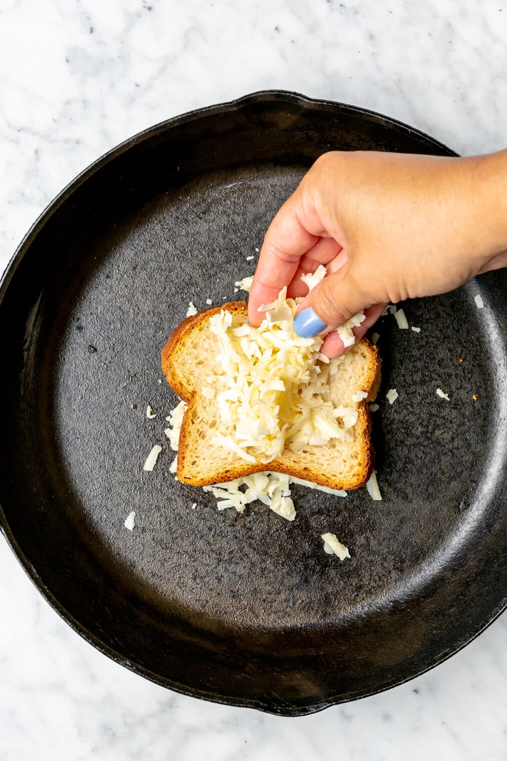 Hand sprinkling cheese on top of a slice of bread in a cast iron pan.