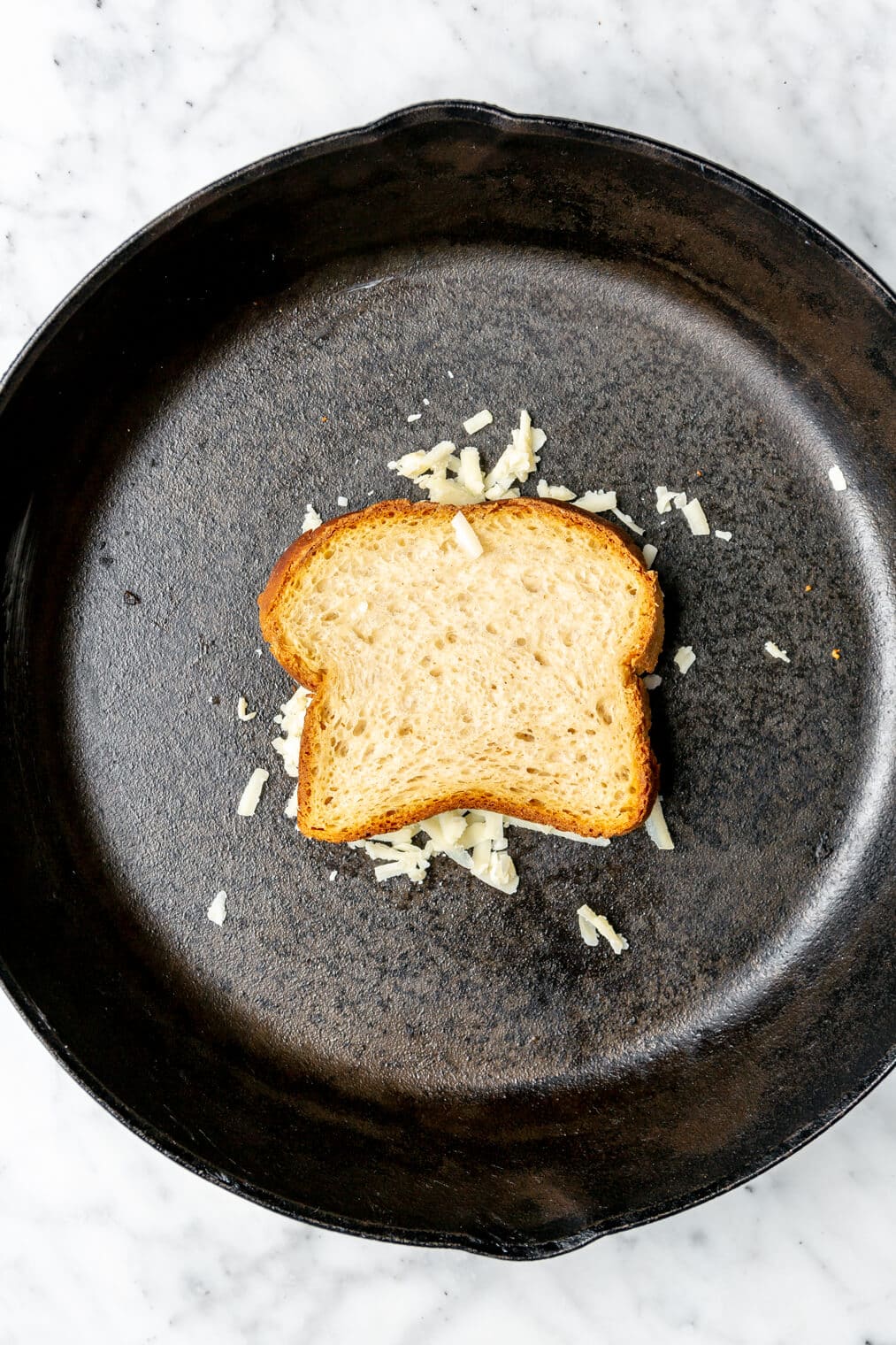 Slice of bread on top of shredded cheese in a cast iron pan.