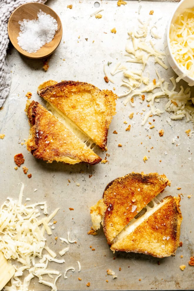 Top down view of two grilled cheese sandwiches cut diagonally in half on a metal sheet pan with shredded cheese sprinkled around the pan and a small wooden bowl of flaky sea salt in the corner.