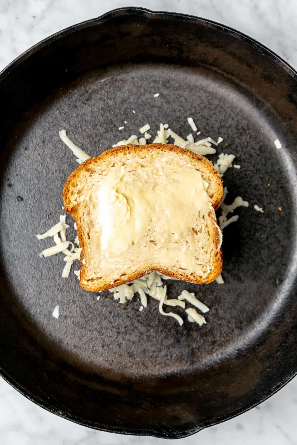 Buttered slice of bread on top of shredded cheese in a cast iron pan.