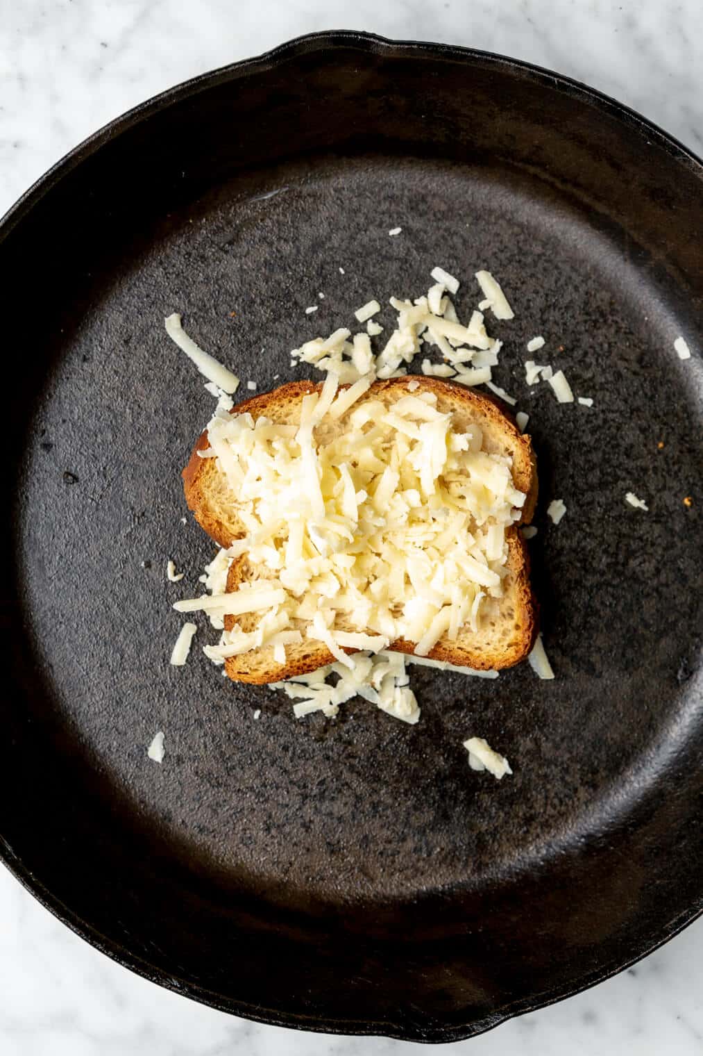 Shredded cheese on top of two slices of bread in cast iron pan.