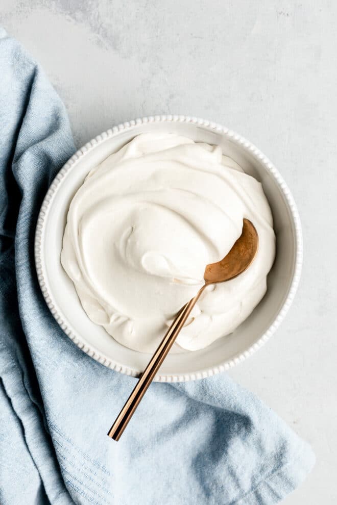 Top down of a bowl of coconut whipped cream with a bronze spoon inside and a light blue linen draped to the left side of the bowl.