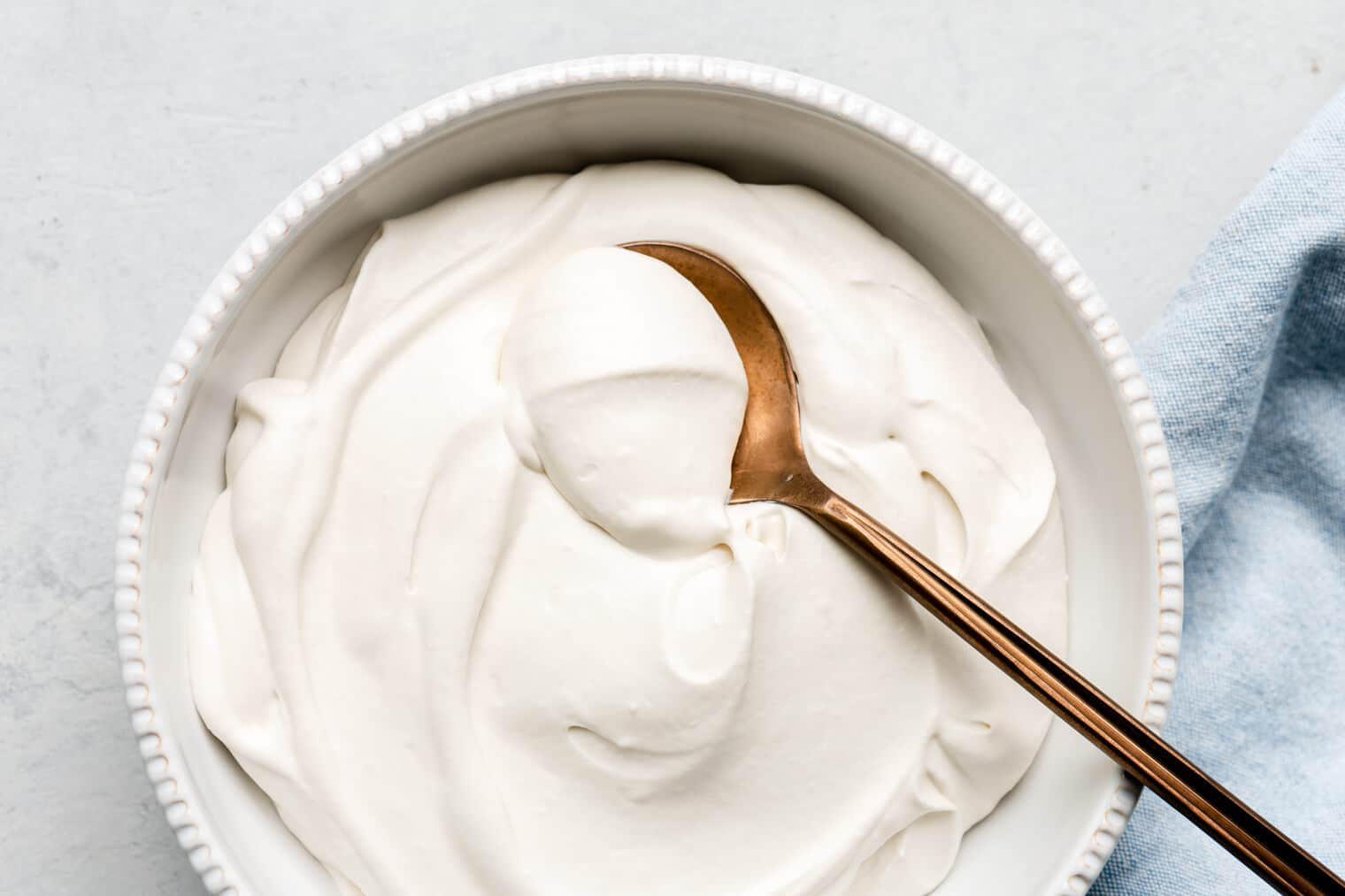 White bowl of coconut whipped cream with a bronze spoon scooping a dollop of whipped coconut cream and a light blue linen draped to the right side.