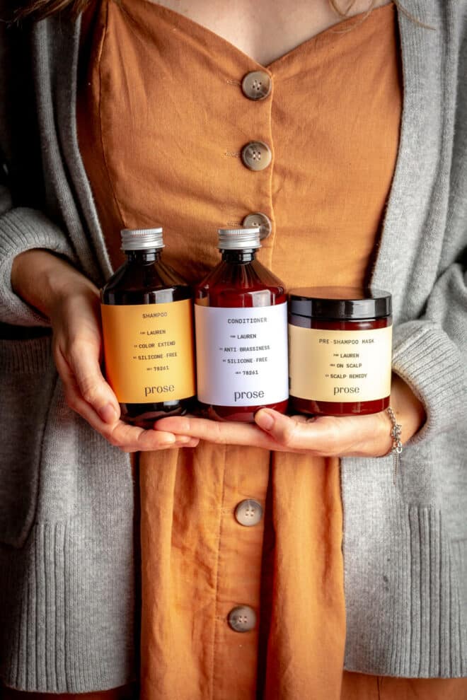 Woman in a burnt orange dress and gray sweater holding Prose hair care system in her hands in front of her waist.