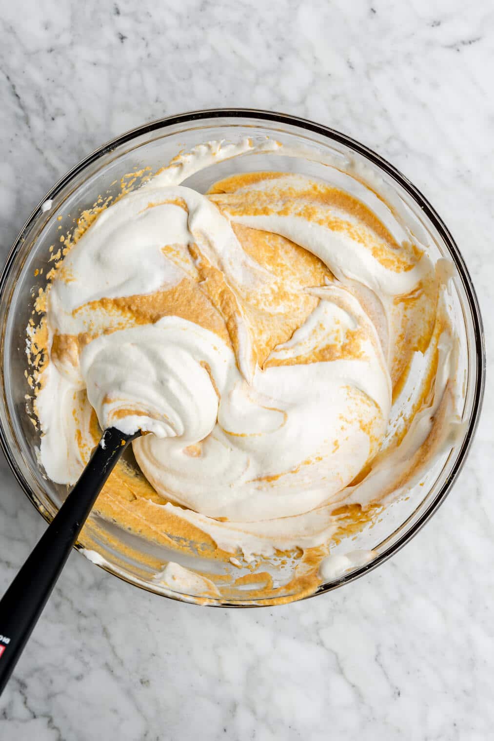 Pumpkin mousse ingredients being folded in with whipped cream in a glass mixing bowl with a black spatula.