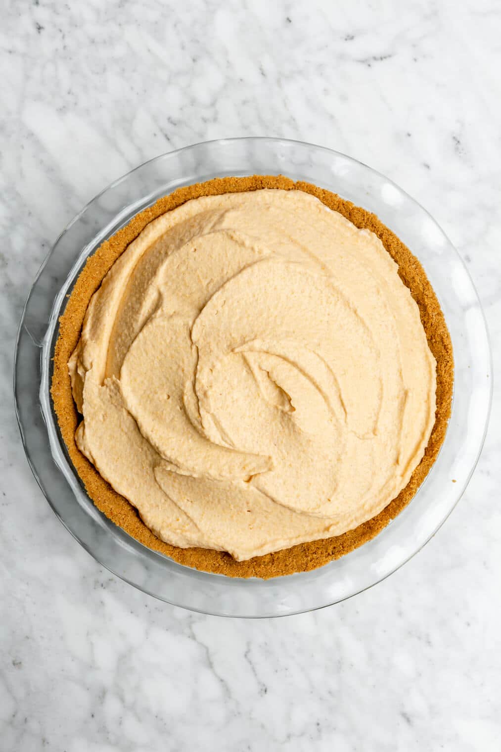 Pumpkin pie mousse with a graham cracker crust in a glass pie dish on a grey and white marble surface.