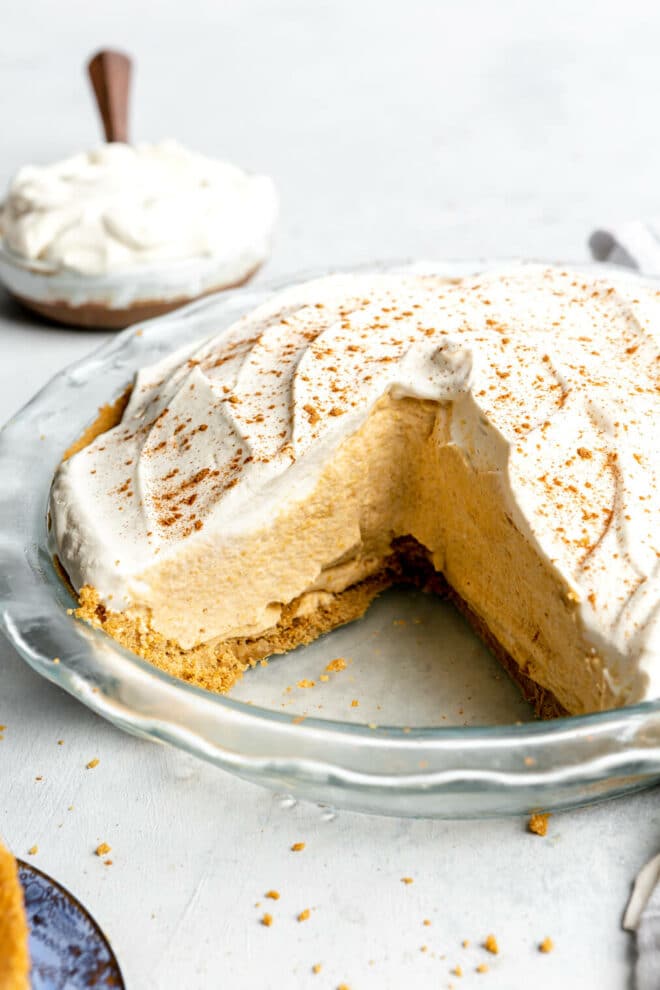 Side view of pumpkin mousse pie in a glass pie dish with one slice removed. There is a layer of graham cracker crust topped with pumpkin mousse and a topped with whipped cream. There is a bowl of whipped cream in the background.