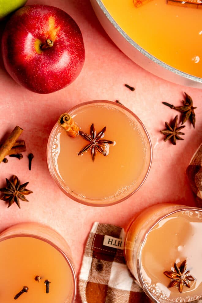 Top down view of glasses with spiced apple cider garnished with star anise on a blush surface with apples and a checkered linen.