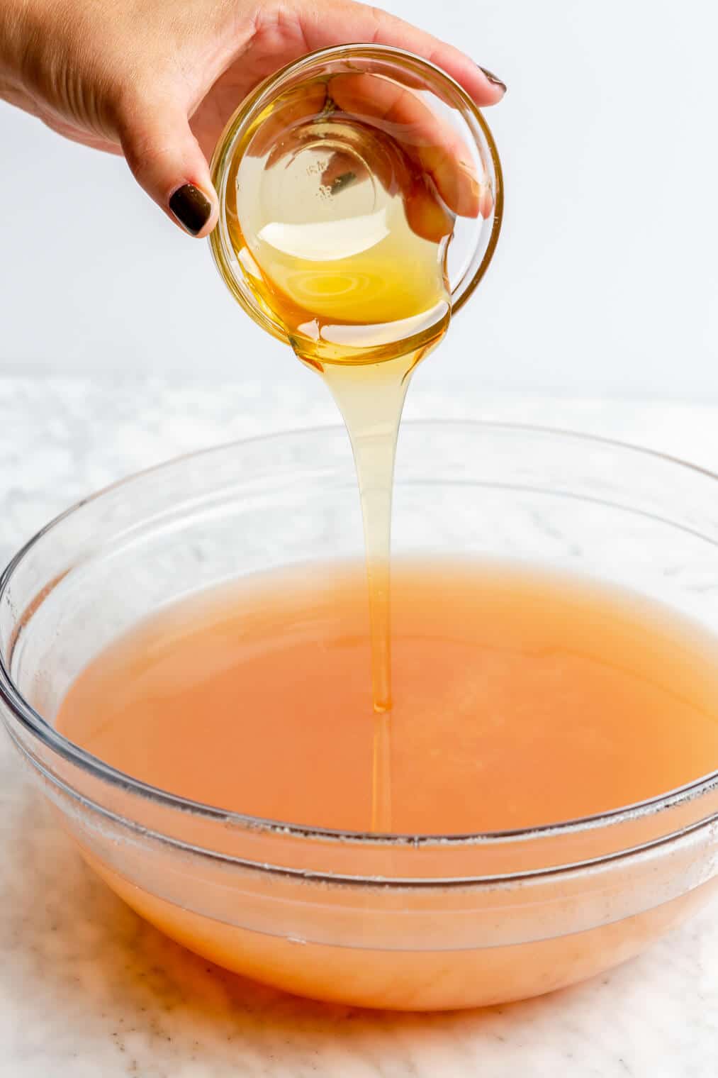 Hand pouring honey into spiced cider liquid in a large glass bowl.