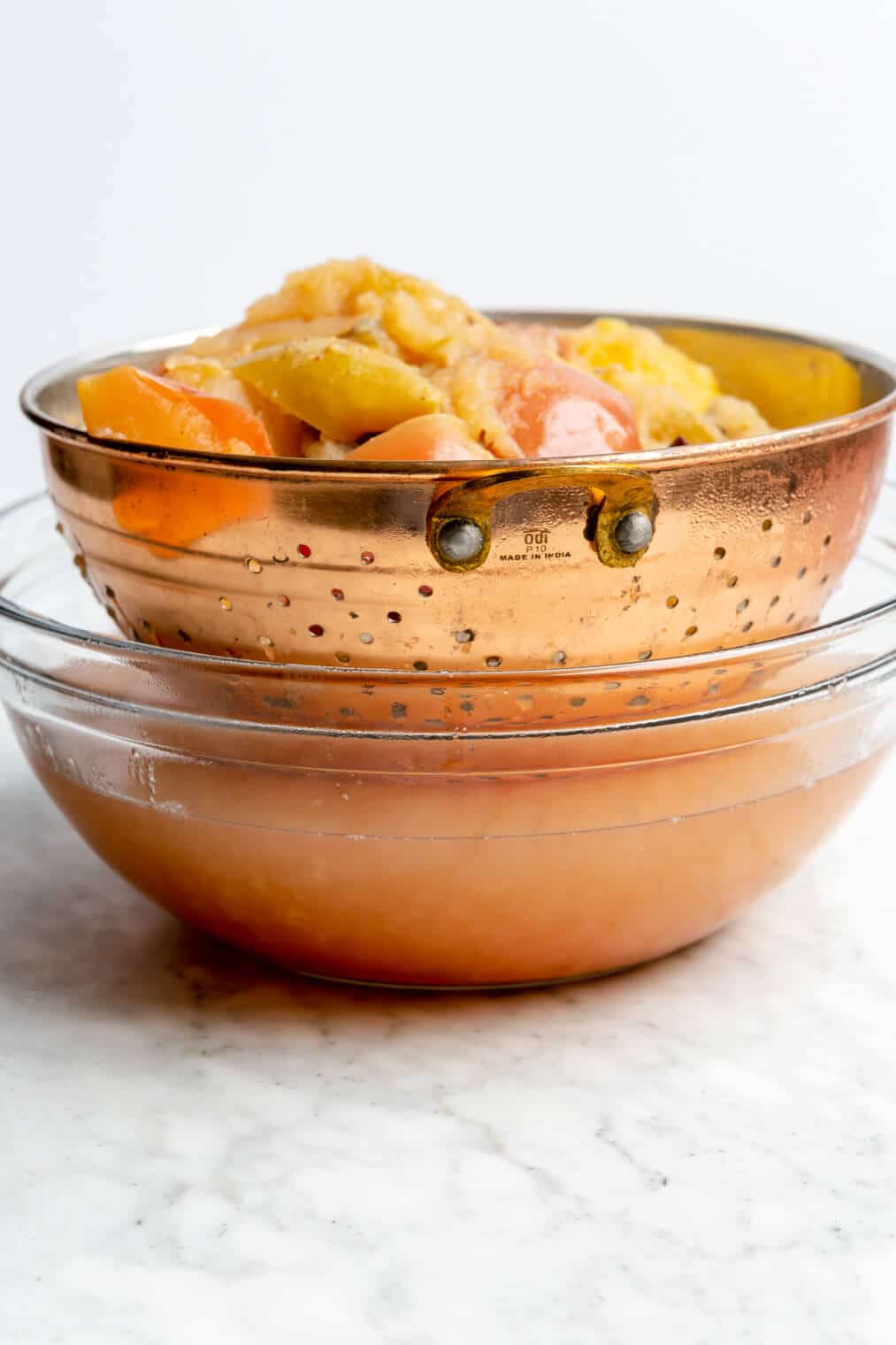 Side view of a strainer with cooked apples and spices and liquid in a large glass bowl.