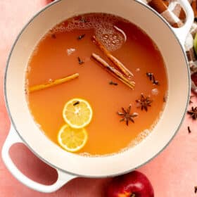 Top down view of pot of cider with slices of lemons, cinnamon sticks, and star anise on a blush surface with red and green apples and a checkered linen.