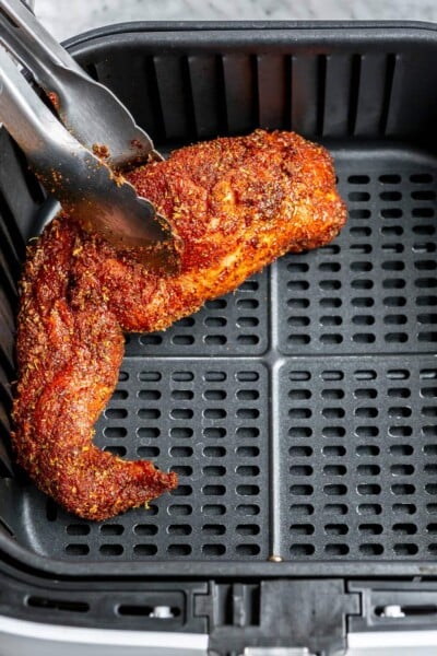 Spiced pork tenderloin being placed into an air fryer basket with a pair of tongs.