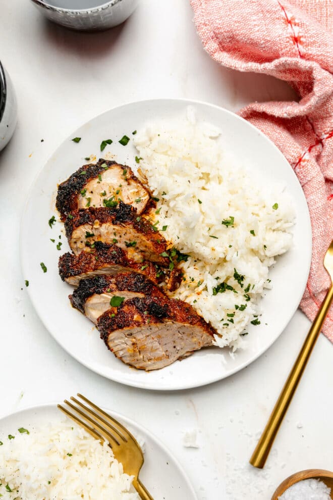White plate with sliced pork tenderloin and a side of rice sprinkled with green herbs on a grey and white marble surface with gold silverware with a red and white striped linen draped in the corner.