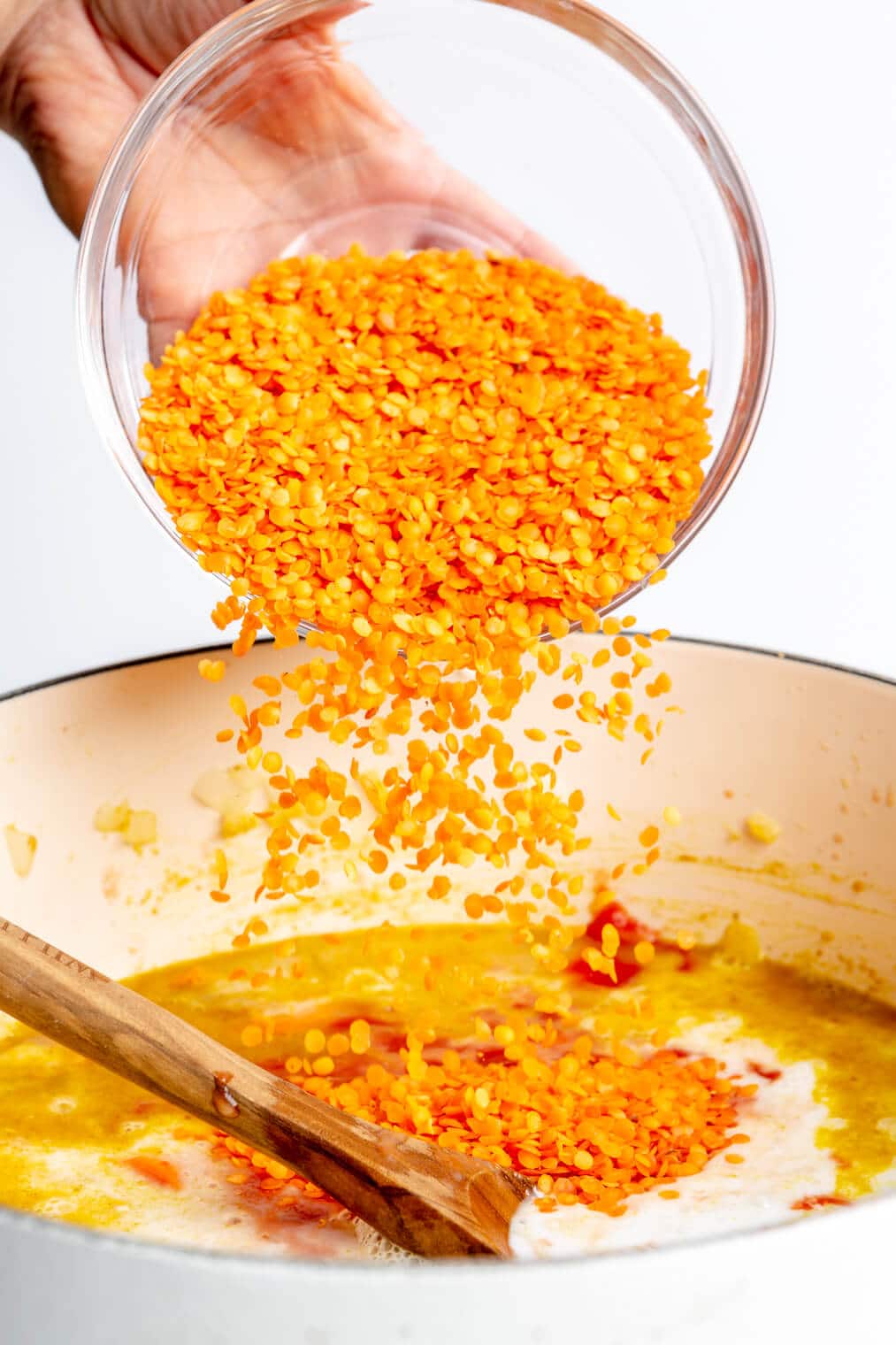 Hand pouring bowl of red lentils into a white pot with a wooden spoon and lentil soup ingredients.