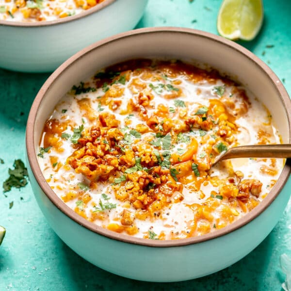 Red lentil soup topped with drizzle of coconut milk, chopped cilantro, and garlic in a white bowl with bronze rim on a turquoise surface with a squeezed lime wedge and sprinkles of chopped cilantro.