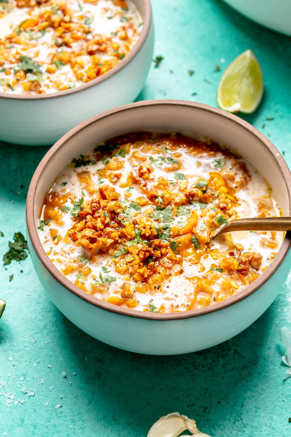 Red lentil soup topped with drizzle of coconut milk, chopped cilantro, and garlic in a white bowl with bronze rim on a turquoise surface with a squeezed lime wedge and sprinkles of chopped cilantro.