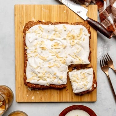 Oatmeal breakfast cake with a corner cut out, topped with coconut whipped cream and coconut flakes on a wooden cutting board.