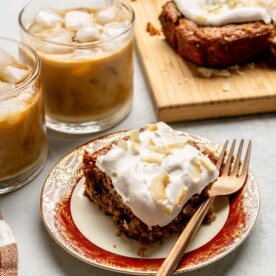 Square piece of oatmeal cake on a white plate with a red and gold decorated rim with a bronze fork and a cutting board with oatmeal cake and two cups of iced coffee in the background.
