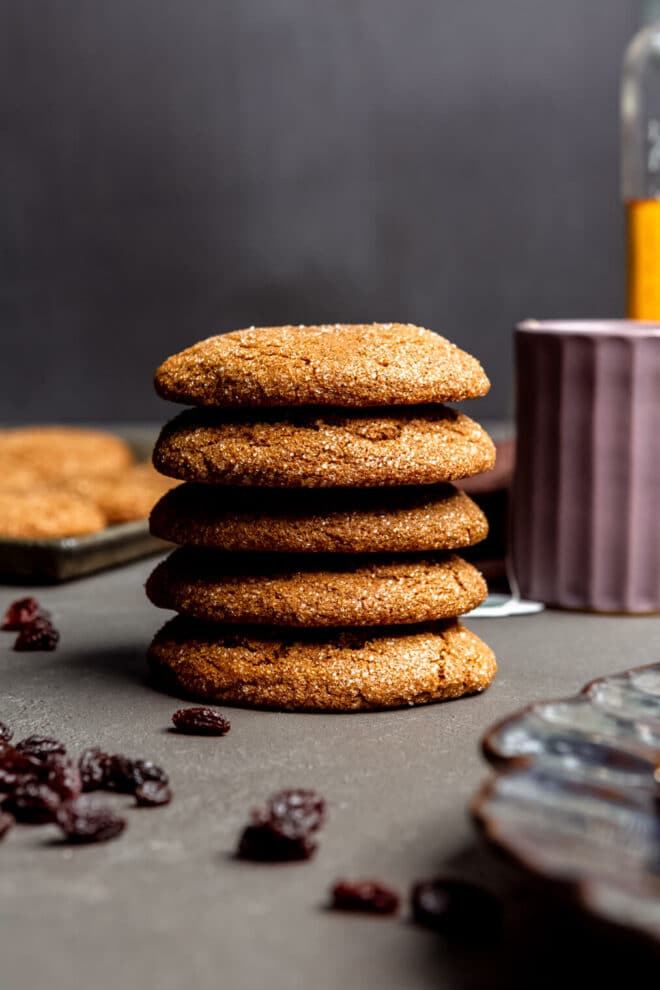 Stack of rum raisin cookies on a slate surface with raisins sprinkled on the table with a plate of cookies and purple mug in the background.