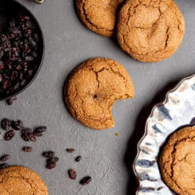 Top down view of rum raisin cookies on a dark slate surface, one cookie with a bite taken out of it, next to a bowl of raisins with a few raisins sprinkled on the table and a shiny, black plate with cookies on the plate.