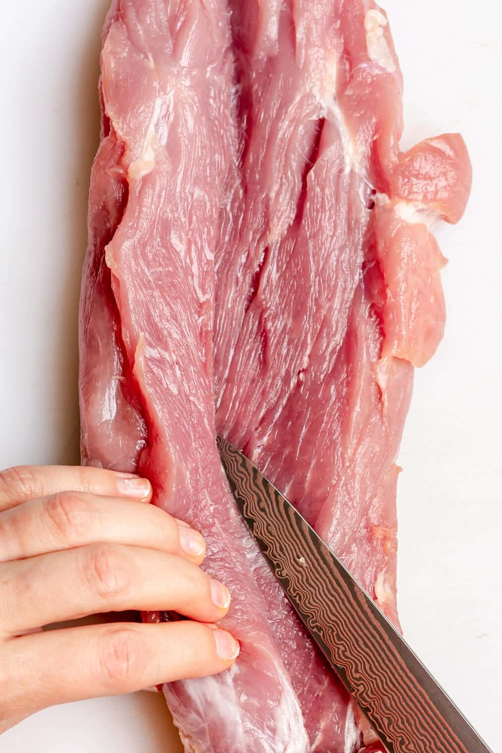 Hand holding pork tenderloin and knife slicing through the middle of the pork tenderloin lengthwise.