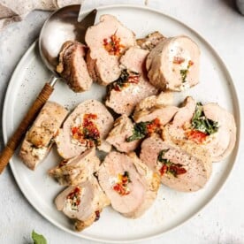 Stuffed pork tenderloin slices on a plate next to a serving spoon on a light grey/white surface.