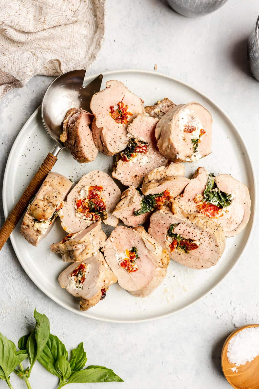 Stuffed pork tenderloin slices on a plate next to a serving spoon on a light grey/white surface.