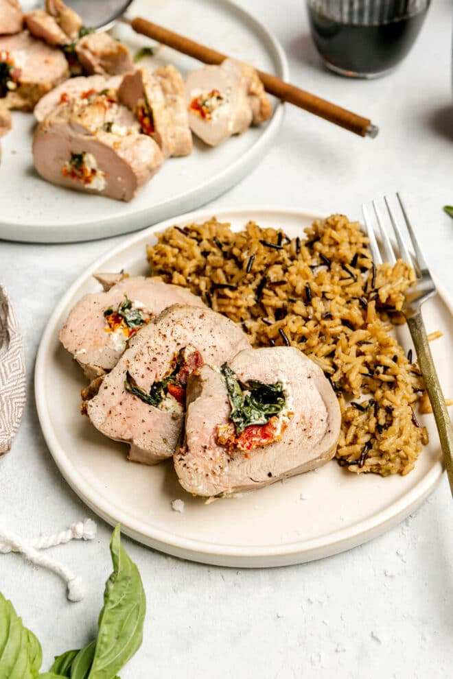 Plate of 3 slices of stuffed pork tenderloin and a side of wild rice with a silver fork. Another plate with tenderloin can be seen in the background. 