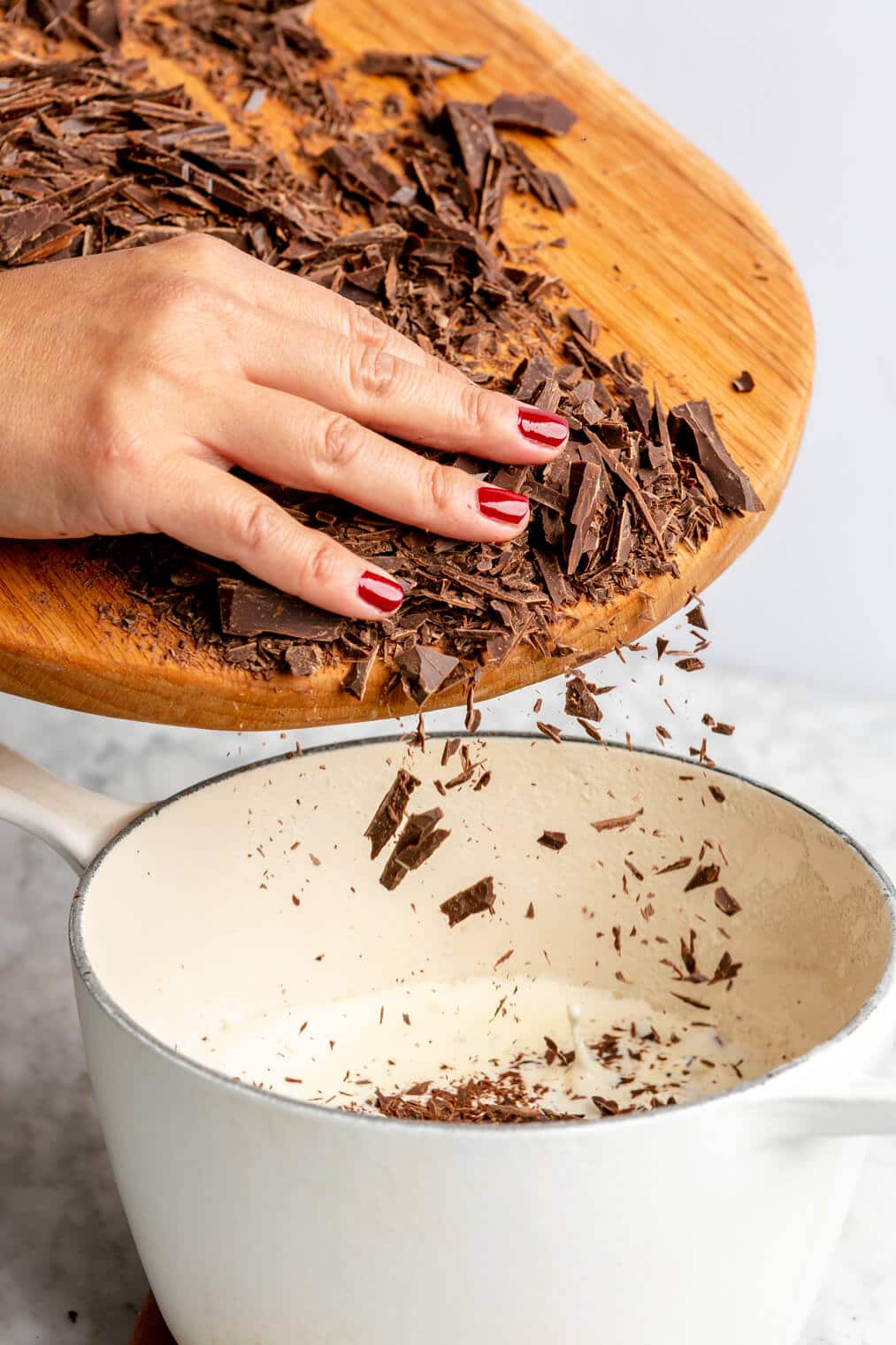 Hand adding shaved chocolate into a white sauce pan with heavy cream from a wooden cutting board.