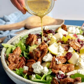 Hand pouring salad dressing over the top of a mixed green salad garnished with goat cheese crumbles, diced pears, candied pecans, and dried cranberries.