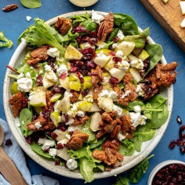 Mixed green salad garnished with goat cheese, candied pecans, pears, and dried cranberries in a large, white bowl on a blue table surrounded by small bowls of garnish, diced pears, salad dressing, and salad servers.
